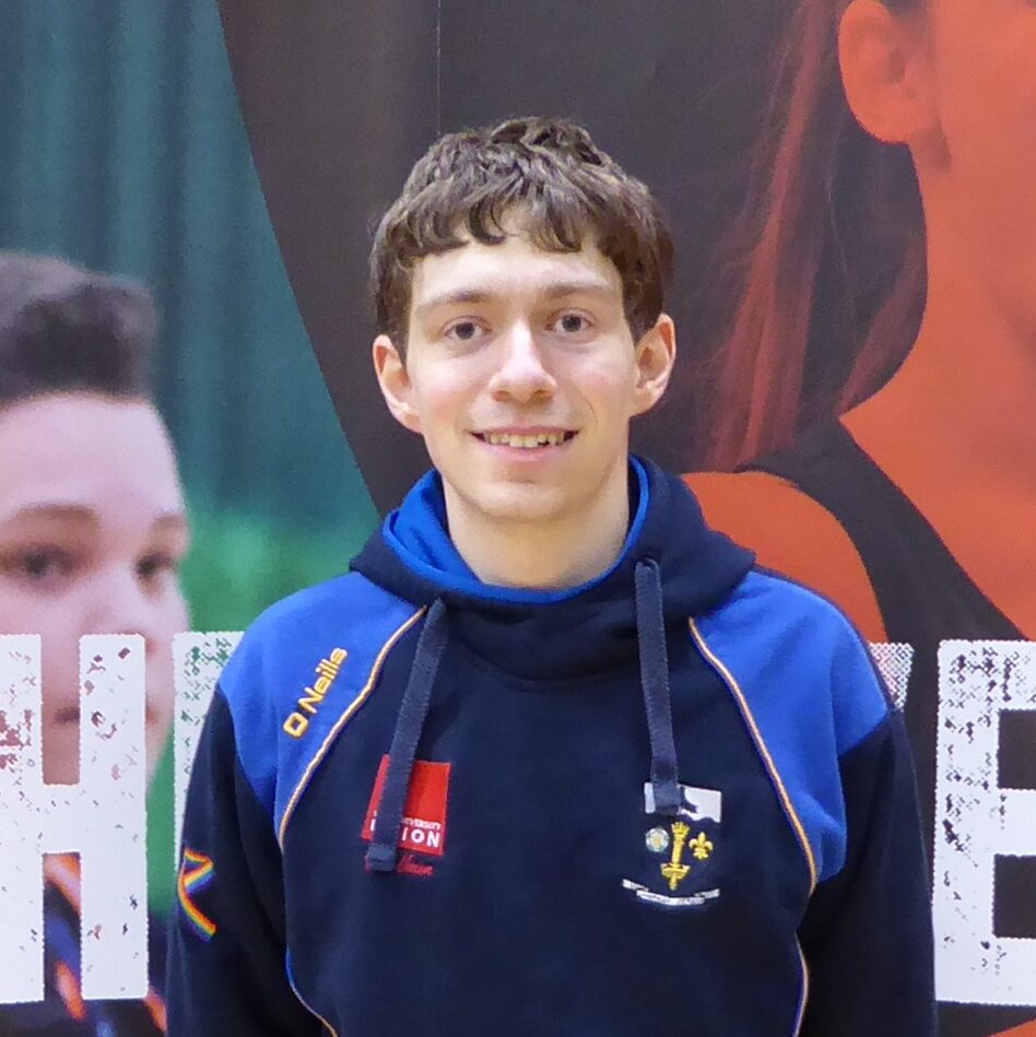James has brown hair, blue eyes and is smiling. He is wearing a navy blue hoodie with blue shoulders